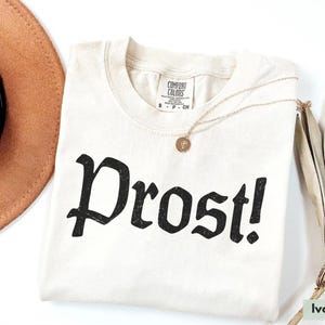 Puede incluir: Camiseta de color marfil con un gr&aacute;fico negro que dice "Prost!" en una fuente g&oacute;tica.