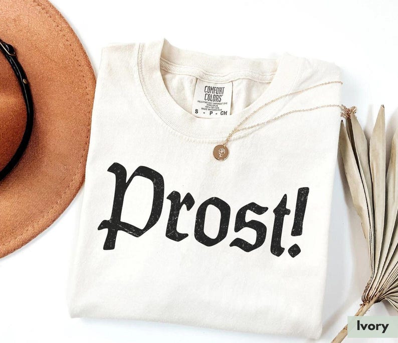 Puede incluir: Camiseta de color marfil con un gr&aacute;fico negro que dice "Prost!" en una fuente g&oacute;tica.