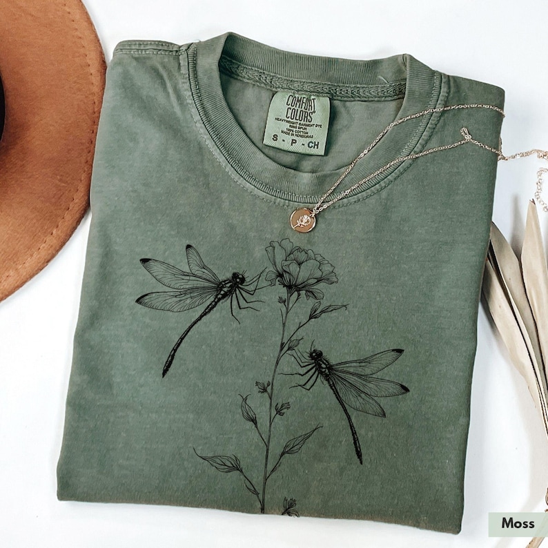 May include: A green t-shirt with a black dragonfly and floral design. The shirt has the text "Comfort Colors" and "S - P - CH" printed on the tag.