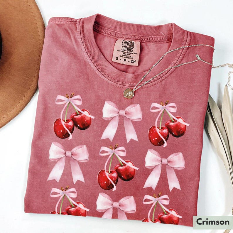 May include: Crimson red t-shirt with a repeating pattern of pink bows and red cherries. The shirt has a crew neck and short sleeves. The text "COMFORT COLORS" is visible on the tag.