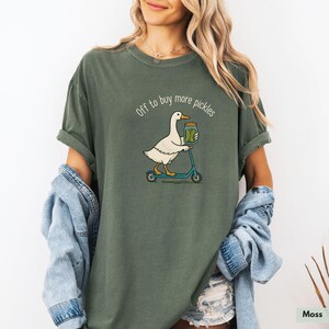 May include: A moss green t-shirt featuring a cartoon goose on a blue scooter, carrying a jar of pickles. The text "Off to buy more pickles" is above the goose. The shirt has a relaxed fit and is made of soft material.