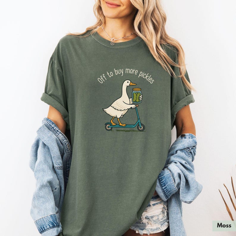 May include: A moss green t-shirt featuring a cartoon goose on a blue scooter, carrying a jar of pickles. The text "Off to buy more pickles" is above the goose. The shirt has a relaxed fit and is made of soft material.