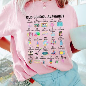 May include: Pink t-shirt with a graphic of an alphabet with old school items like a boom box, cassette tape, and a hula hoop. The text reads "OLD SCHOOL ALPHABET".