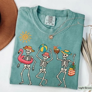 May include: Light green t-shirt with a tie-dye effect featuring a graphic of four skeletons dressed in summer attire. The skeletons are holding a flamingo float, a beach ball, a tropical drink, and a beach bag. The shirt has the text "Comfort Colors" and "S - P - CH" printed on the front.