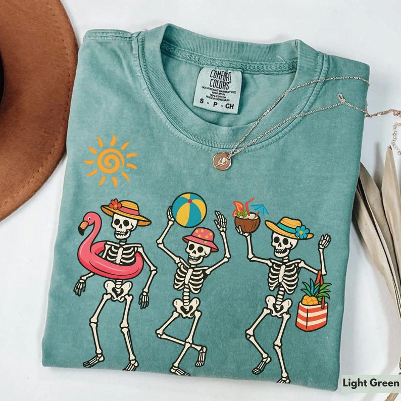 May include: Light green t-shirt with a tie-dye effect featuring a graphic of four skeletons dressed in summer attire. The skeletons are holding a flamingo float, a beach ball, a tropical drink, and a beach bag. The shirt has the text "Comfort Colors" and "S - P - CH" printed on the front.