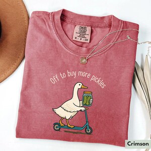 May include: Crimson t-shirt featuring the phrase "Off to buy more pickles" and a cartoon duck on a scooter with a pickle jar. The shirt has a crew neck and a small tag. The colour is described as crimson.