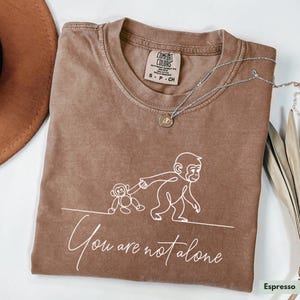 May include: Espresso-colored t-shirt with a white line drawing of a monkey holding the hand of a smaller monkey. The text "You are not alone" is below the drawing. A silver necklace with a pendant is on the shirt.