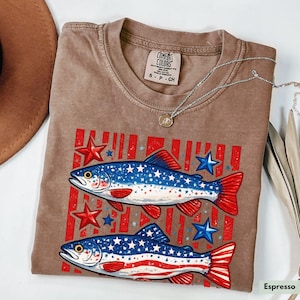 May include: Espresso-colored t-shirt featuring a graphic of two fish with red and blue star and stripe designs. The shirt has a crew neck and short sleeves. A silver necklace with a small pendant is draped across the shirt.