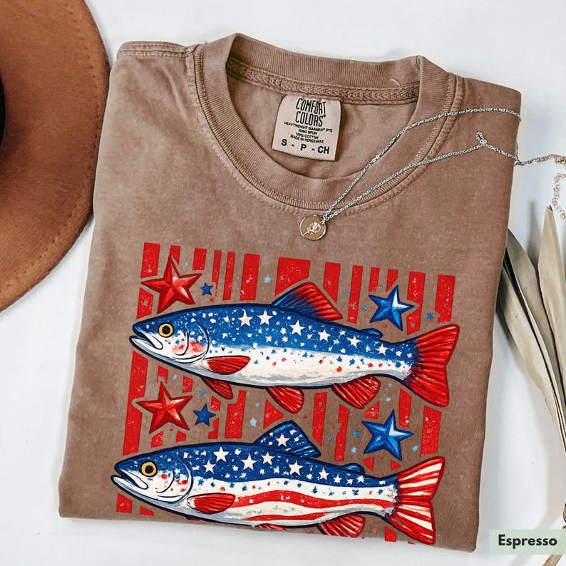May include: Espresso-colored t-shirt featuring a graphic of two fish with red and blue star and stripe designs. The shirt has a crew neck and short sleeves. A silver necklace with a small pendant is draped across the shirt.