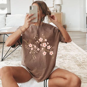 May include: A brown t-shirt with a floral print of pink cherry blossoms. The t-shirt has a relaxed fit and short sleeves.