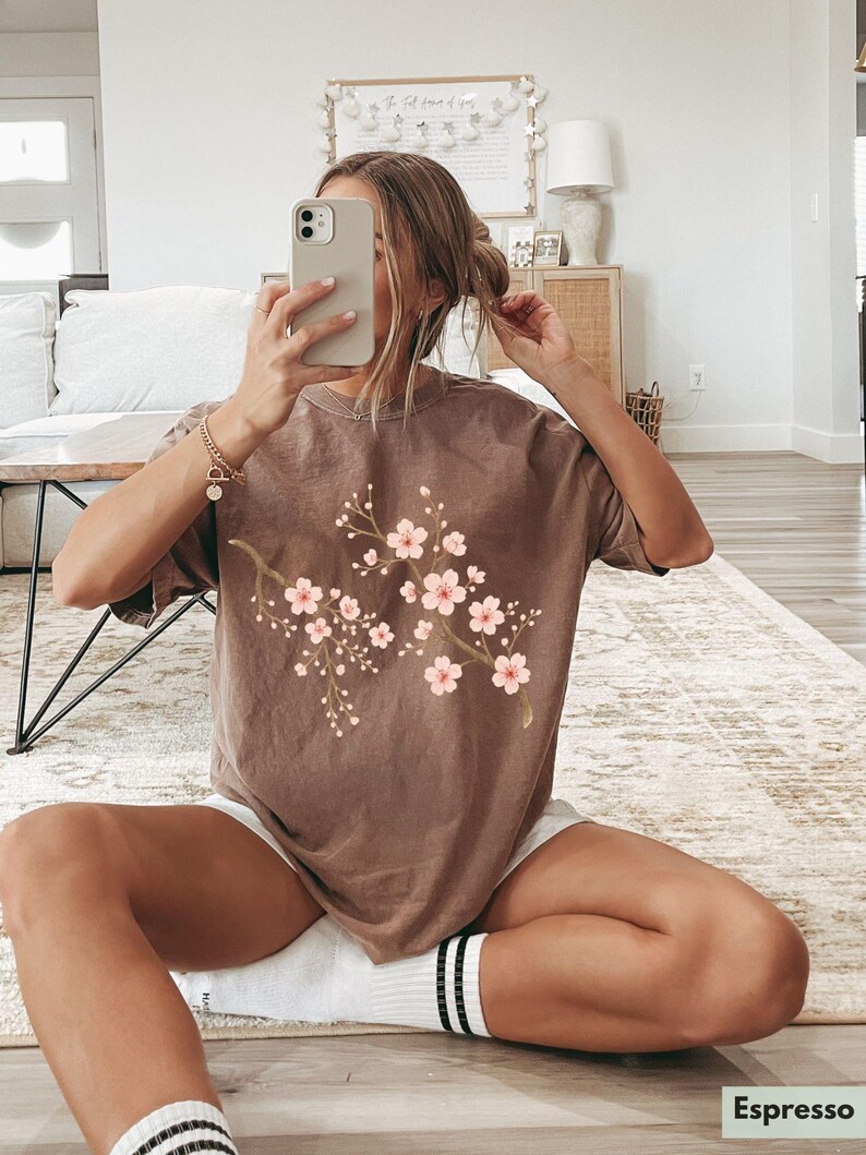 May include: A brown t-shirt with a floral print of pink cherry blossoms. The t-shirt has a relaxed fit and short sleeves.