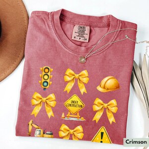 May include: Crimson red t-shirt with yellow construction themed graphics. The graphics include a traffic light, a hard hat, a construction sign that says "Under Construction", and several yellow bows.