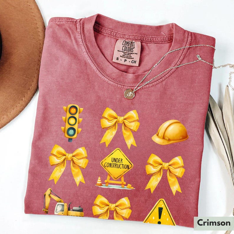 May include: Crimson red t-shirt with yellow construction themed graphics. The graphics include a traffic light, a hard hat, a construction sign that says "Under Construction", and several yellow bows.