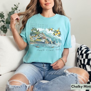 以下が含まれることがあります： イタリアのアマルフィ海岸のグラフィックプリントが施された薄いブルーのTシャツ。グラフィックには、カラフルな街並みとレモンの木、そして「Amalfi Coast, Italy」の文字が描かれています。