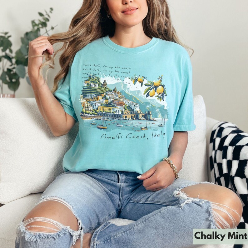以下が含まれることがあります： イタリアのアマルフィ海岸のグラフィックプリントが施された薄いブルーのTシャツ。グラフィックには、カラフルな街並みとレモンの木、そして「Amalfi Coast, Italy」の文字が描かれています。