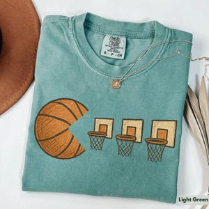 Op de afbeelding: Lichtgroen Comfort Colors T-shirt met een basketbalthema. Het ontwerp toont een basketbal met een Pac-Man-achtige hap eruit en drie basketbalringen. Een zilveren ketting met een hanger is over het shirt gedrapeerd.