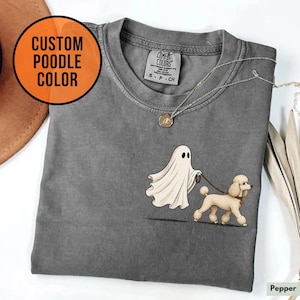 May include: A charcoal grey t-shirt featuring a graphic of a white ghost walking a tan poodle on a lead. The shirt has a crew neckline and a Comfort Colors brand label inside the collar. A silver chain necklace rests on the fabric near the neckline.