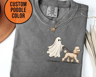 Puede incluir: Una camiseta gris carbón con un gráfico de un fantasma blanco paseando a un caniche color canela con una correa. La camiseta tiene cuello redondo y una etiqueta de la marca Comfort Colors. Una cadena plateada descansa sobre la tela.