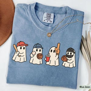 May include: Blue jean colored t-shirt with a graphic of four cartoon ghosts playing baseball. Each ghost wears a baseball cap or helmet and holds a bat, ball, drink, or foam finger. The shirt has a Comfort Colors tag.