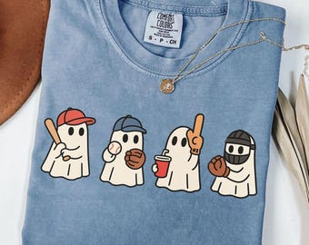 May include: Blue jean colored t-shirt with a graphic of four cartoon ghosts playing baseball. Each ghost wears a baseball cap or helmet and holds a bat, ball, drink, or foam finger. The shirt has a Comfort Colors tag.