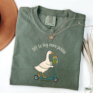 May include: A moss green Comfort Colors t-shirt featuring the text "Off to buy more pickles" and a cartoon duck on a scooter with a pickle jar. A silver necklace is draped across the shirt.
