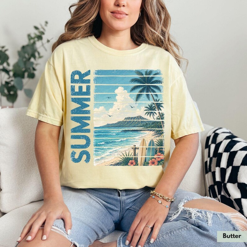 May include: Butter-colored t-shirt featuring a tropical beach scene with palm trees, ocean waves, and the word "SUMMER" in blue vertical text. The graphic includes surfboards and flowers, evoking a vacation theme.