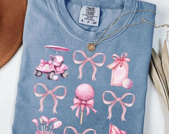 May include: A light blue t-shirt with a pink golf cart, golf balls, golf clubs, and pink bows. The shirt has the text "Comfort Colors" on the tag.