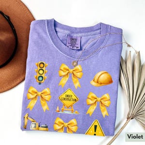 May include: A purple t-shirt with yellow construction-themed graphics, including a traffic light, a hard hat, a construction sign that says "Under Construction", and bows. The shirt is made by Comfort Colors.