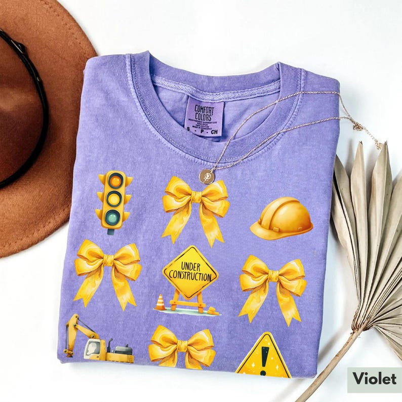 May include: A purple t-shirt with yellow construction-themed graphics, including a traffic light, a hard hat, a construction sign that says "Under Construction", and bows. The shirt is made by Comfort Colors.