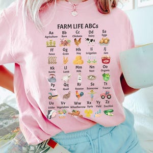 May include: A pink t-shirt featuring a graphic of farm-related items and the alphabet. Each letter is paired with a farm-related word and a small illustration. The text reads "FARM LIFE ABCS".