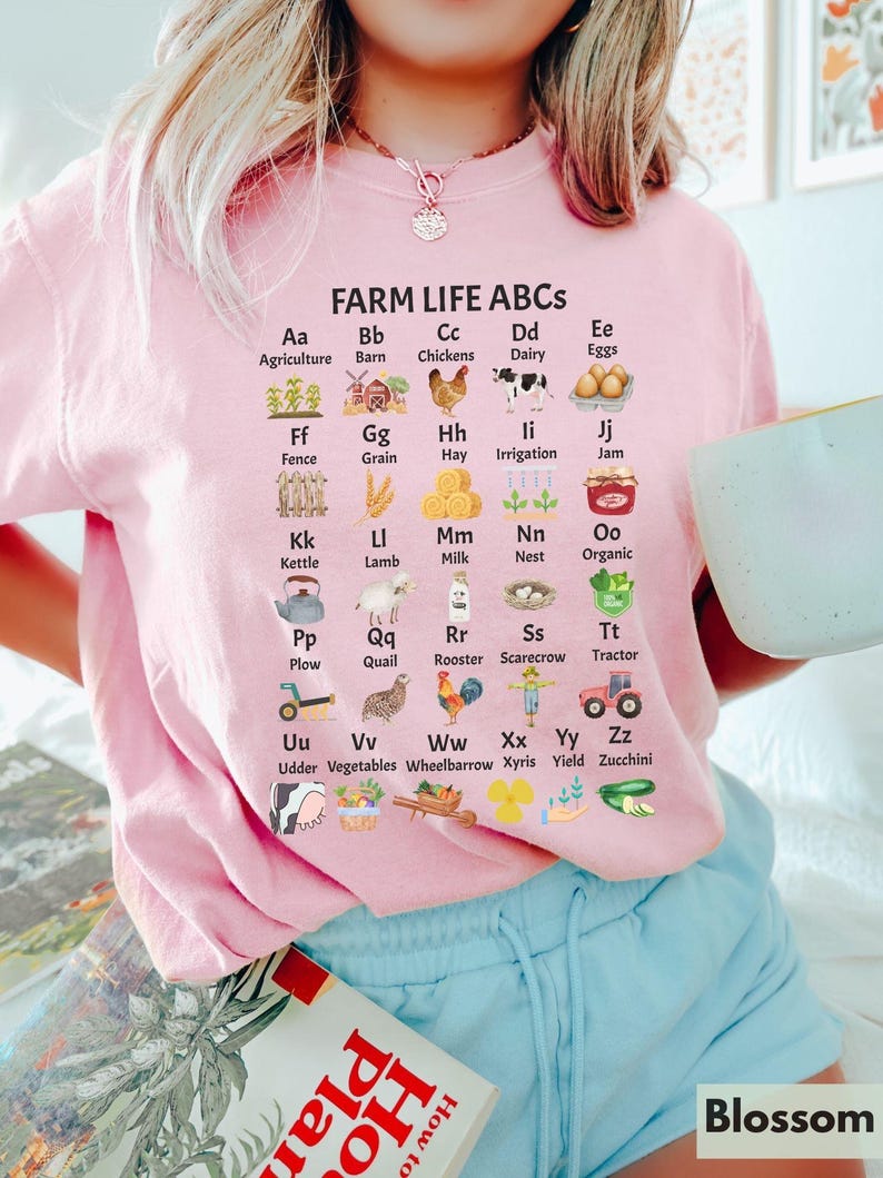 May include: A pink t-shirt featuring a graphic of farm-related items and the alphabet. Each letter is paired with a farm-related word and a small illustration. The text reads "FARM LIFE ABCS".
