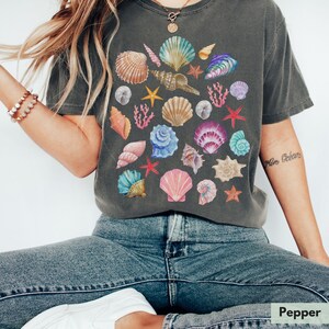 Op de afbeelding: Een donkergrijs T-shirt met een kleurrijk schelpmotief. Het shirt heeft een losse pasvorm en een ronde hals. De tekst "More Ocean" is gedrukt op de rechtermouw.