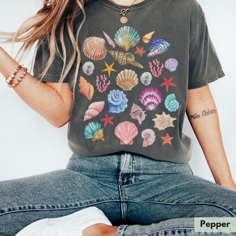 Op de afbeelding: Een donkergrijs T-shirt met een kleurrijk schelpmotief. Het shirt heeft een losse pasvorm en een ronde hals. De tekst "More Ocean" is gedrukt op de rechtermouw.