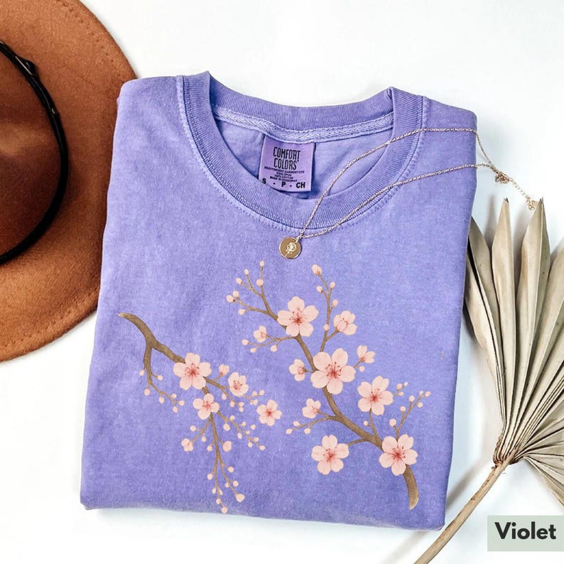 May include: A purple t-shirt with a floral design featuring two branches of pink cherry blossoms. The shirt has a tag that reads "Comfort Colors".