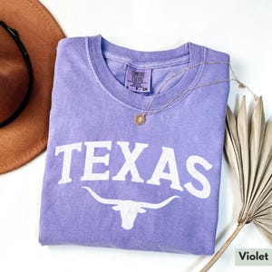 Puede incluir: Una camiseta morada con la palabra "TEXAS" en letras blancas y una silueta blanca de la cabeza de un toro longhorn con cuernos. La camiseta est&aacute; doblada y colocada sobre una superficie blanca.