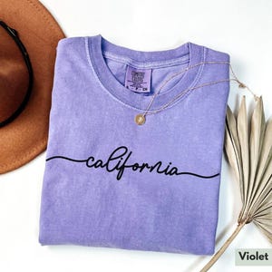 May include: A purple t-shirt with the word "california" written in a cursive font. The shirt is made of a soft, comfortable material and has a relaxed fit.