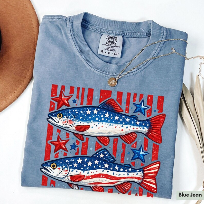 May include: Blue jean colored t-shirt featuring two fish with red, white, and blue patriotic designs. The fish are set against a background of red stripes and stars. The shirt has a crew neck and short sleeves. A gold necklace is draped across the shirt.