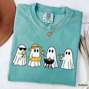 May include: Seafoam green Comfort Colors t-shirt featuring a graphic of four cartoon ghosts enjoying summer activities. One ghost wears sunglasses and eats ice cream, another wears a hat and floatie, one grills, and the last one sips a drink.