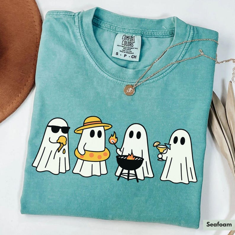 May include: Seafoam green Comfort Colors t-shirt featuring a graphic of four cartoon ghosts enjoying summer activities. One ghost wears sunglasses and eats ice cream, another wears a hat and floatie, one grills, and the last one sips a drink.