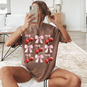 May include: A brown t-shirt with a graphic print of pink bows and red cherries. The shirt is made of a soft, comfortable material.