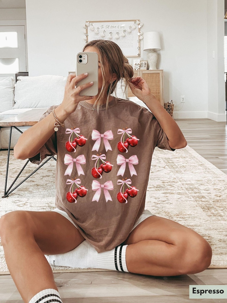 May include: A brown t-shirt with a graphic print of pink bows and red cherries. The shirt is made of a soft, comfortable material.