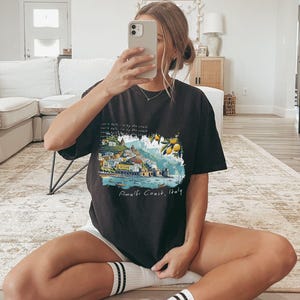 以下が含まれることがあります： イタリアのアマルフィ海岸のグラフィックプリントが施された黒いTシャツ。テキスト「Amalfi Coast, Italy」はグラフィックの下に印刷されています。