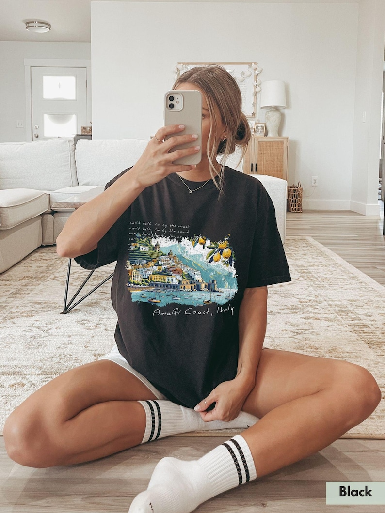 以下が含まれることがあります： イタリアのアマルフィ海岸のグラフィックプリントが施された黒いTシャツ。テキスト「Amalfi Coast, Italy」はグラフィックの下に印刷されています。