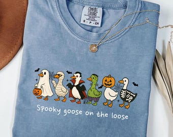 May include: Blue jean colored t-shirt with a Halloween-themed graphic. The graphic features a line of cartoon geese dressed as a ghost, mummy, vampire, Frankenstein, pumpkin, and skeleton. The text "Spooky goose on the loose" is below the geese.