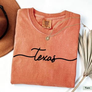 May include: A coral colored t-shirt with the word "Texas" written in a cursive font. The shirt is folded and has a tag that says "Comfort Colors".