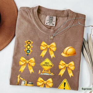 May include: A brown t-shirt with yellow construction-themed graphics. The graphics include a traffic light, a construction sign that says "Under Construction", a hard hat, a bulldozer, a warning sign, and four yellow bows.
