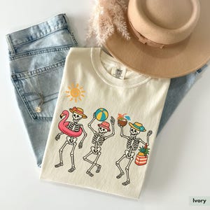 May include: Ivory t-shirt with a graphic of three skeletons wearing summer accessories, including a pink flamingo float, a beach ball, and a pineapple. The skeletons are wearing hats and sunglasses. The graphic is surrounded by a sun and a palm tree.
