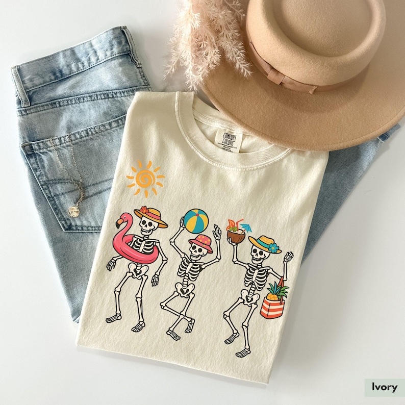 May include: Ivory t-shirt with a graphic of three skeletons wearing summer accessories, including a pink flamingo float, a beach ball, and a pineapple. The skeletons are wearing hats and sunglasses. The graphic is surrounded by a sun and a palm tree.