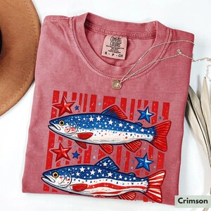 May include: Crimson-colored t-shirt featuring a graphic of two fish with a patriotic design. The fish have blue bodies with white stars and red and white striped tails. Red stars and blue stars are also in the design. The shirt has a crew neck and short sleeves.
