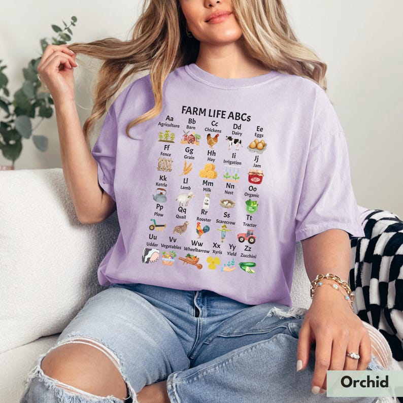 May include: A purple t-shirt with a white graphic of a farm alphabet chart. The chart features illustrations of farm animals, plants, and farm equipment. The text on the chart reads "FARM LIFE ABCS".